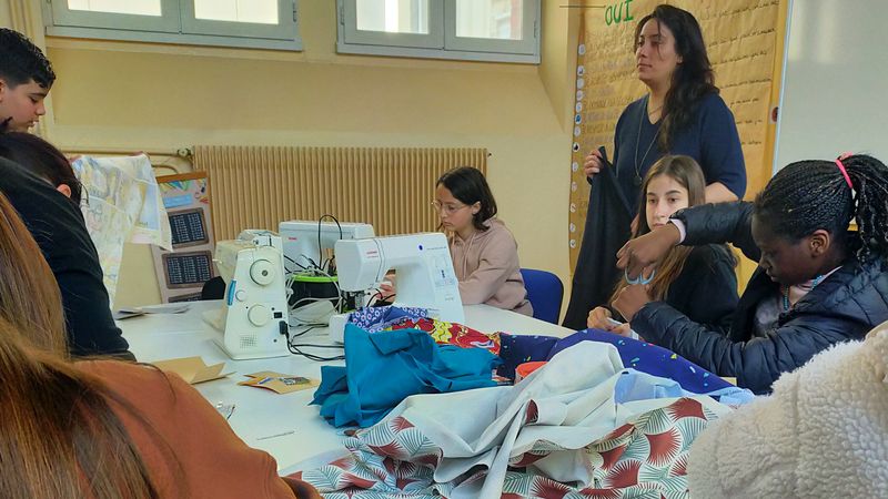 L'image représente des jeunes enfants autour d'une table sur laquelle on peut voir plusieurs machine à coudre et de nombreux morceaux de tissus de couleurs différentes. Les enfants utilisent des machines ou des ciseaux sur le tissu, sous l'oeil de deux adultes. 