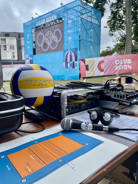 La photo montre en fond l'écran géant du village olympique, et au premier plan du matériel informatique, un micro, un casque, ainsi qu'un ballon de beach volley et une reproduction à petite échelle d'un terrain