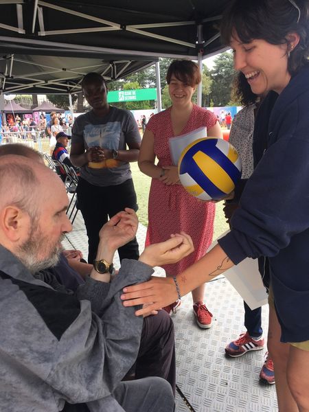 Une personne aveugle tend les mains vers une femme tenant contre un elle un ballon de beach-volley qu'elle s'apprête à lui faire découvrir. L'ensemble des personnes sur la photo sourie. 