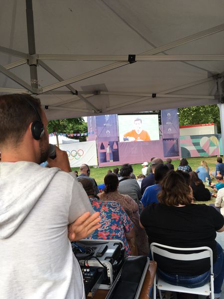 Une personne debout, avec un micro, audiodécrit un match que l'on aperçoit sur le grand écran en fond, sous le regard de plusieurs personnes assises