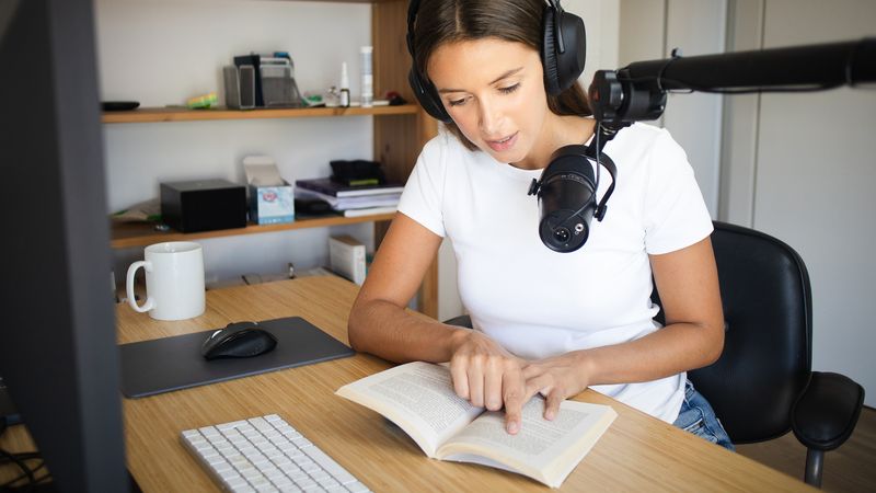 Sur l'image une femme est assises à son bureau et lit un livre devant un micro, avec un casque audio sur les oreilles. 