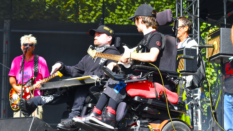 Sur la photo on peut voir deux personnes en fauteuil roulant électrique jouant de la guitare sur une scène de concert ensoleillée