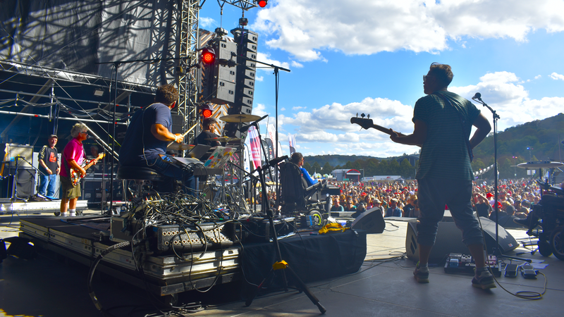 La photo d'un concert vu du fond de la scène, avec un batteur en action, un bassiste et un guitariste vu de derrière, en fond un public nombreux qui danse sous le ciel bleu