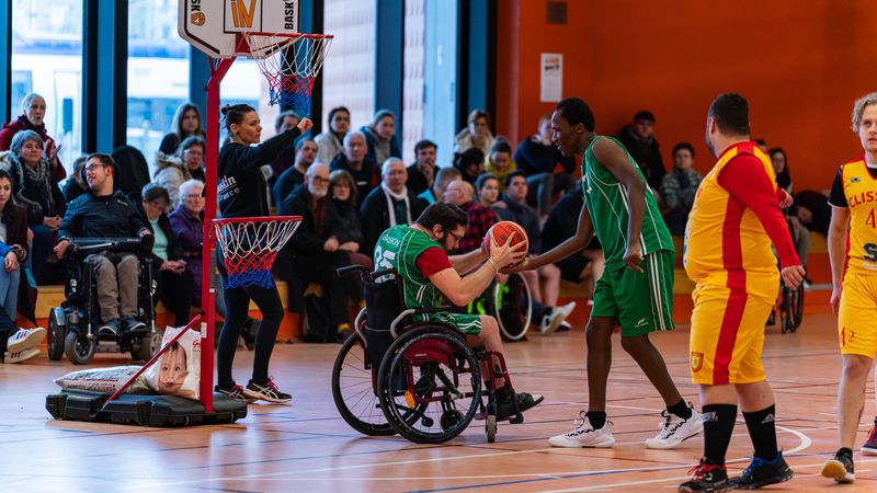 Photo d'un match de baskin ou debant un public nombreux on peut voir deux joueurs d'orea, maillots verts, un en fauteuil roulant donnant le ballon à un autre joueur debout.