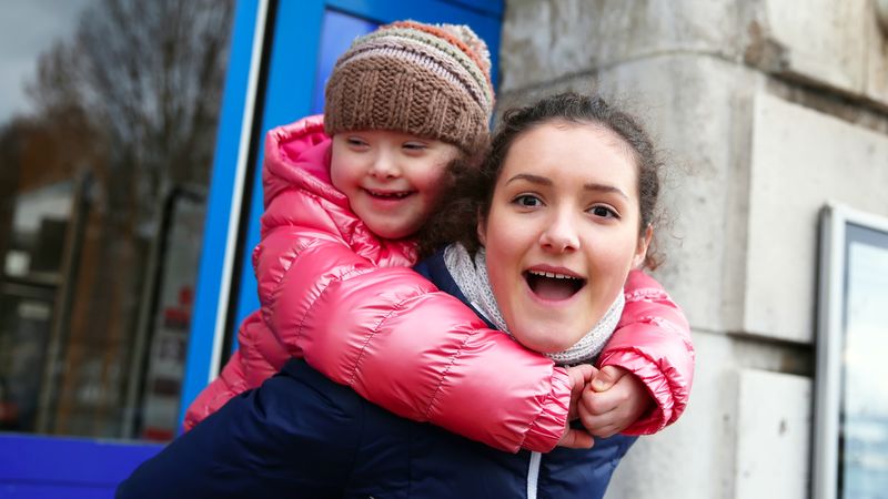 Jeune enfant trisomique sur le dos d'une jeune femme. Les deux affichent un grand sourire. 