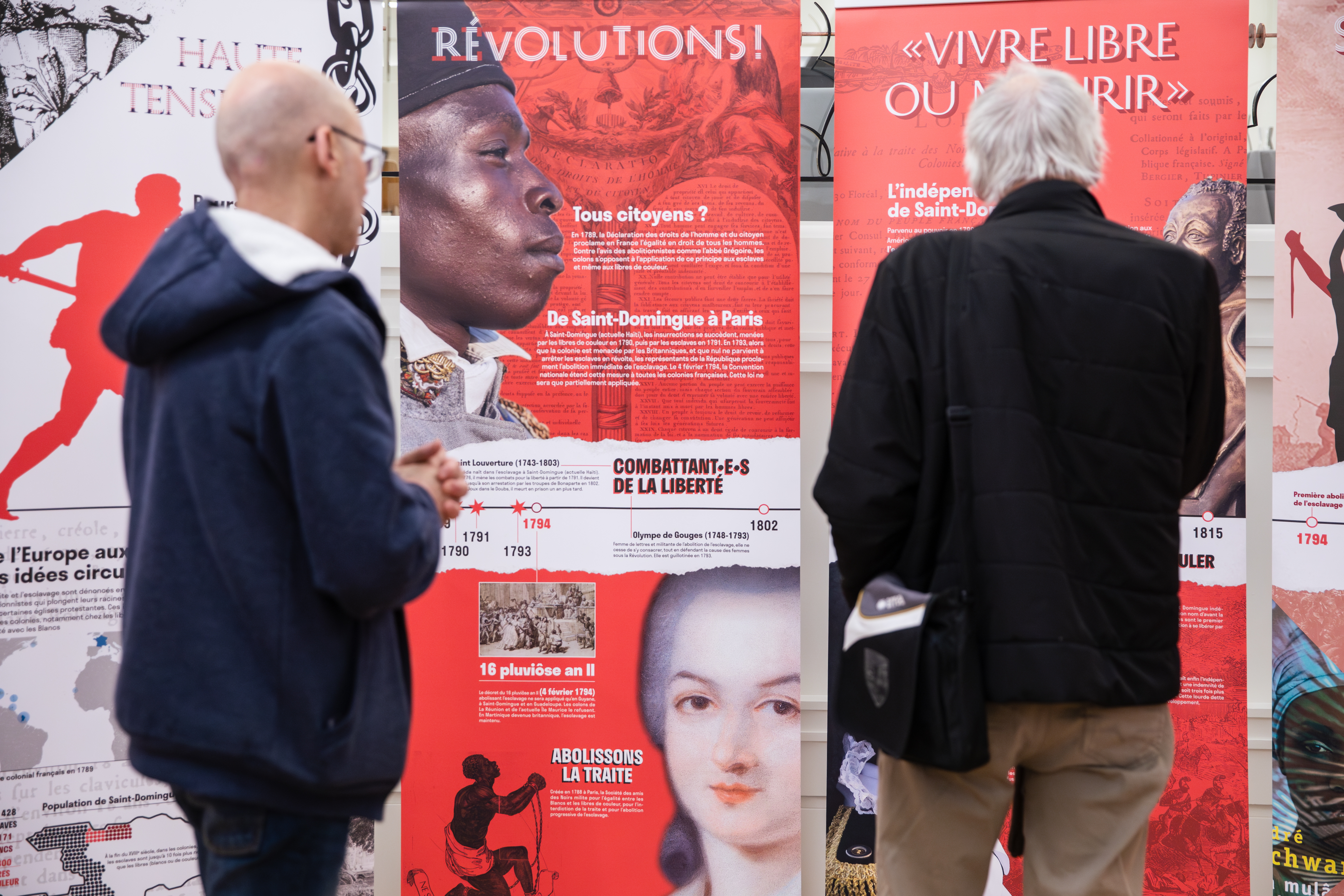 Plusieurs panneaux de couleurs rouge vive présentant des figures historique. Au premier plan, deux hommes sont en train de regarder l'exposition.