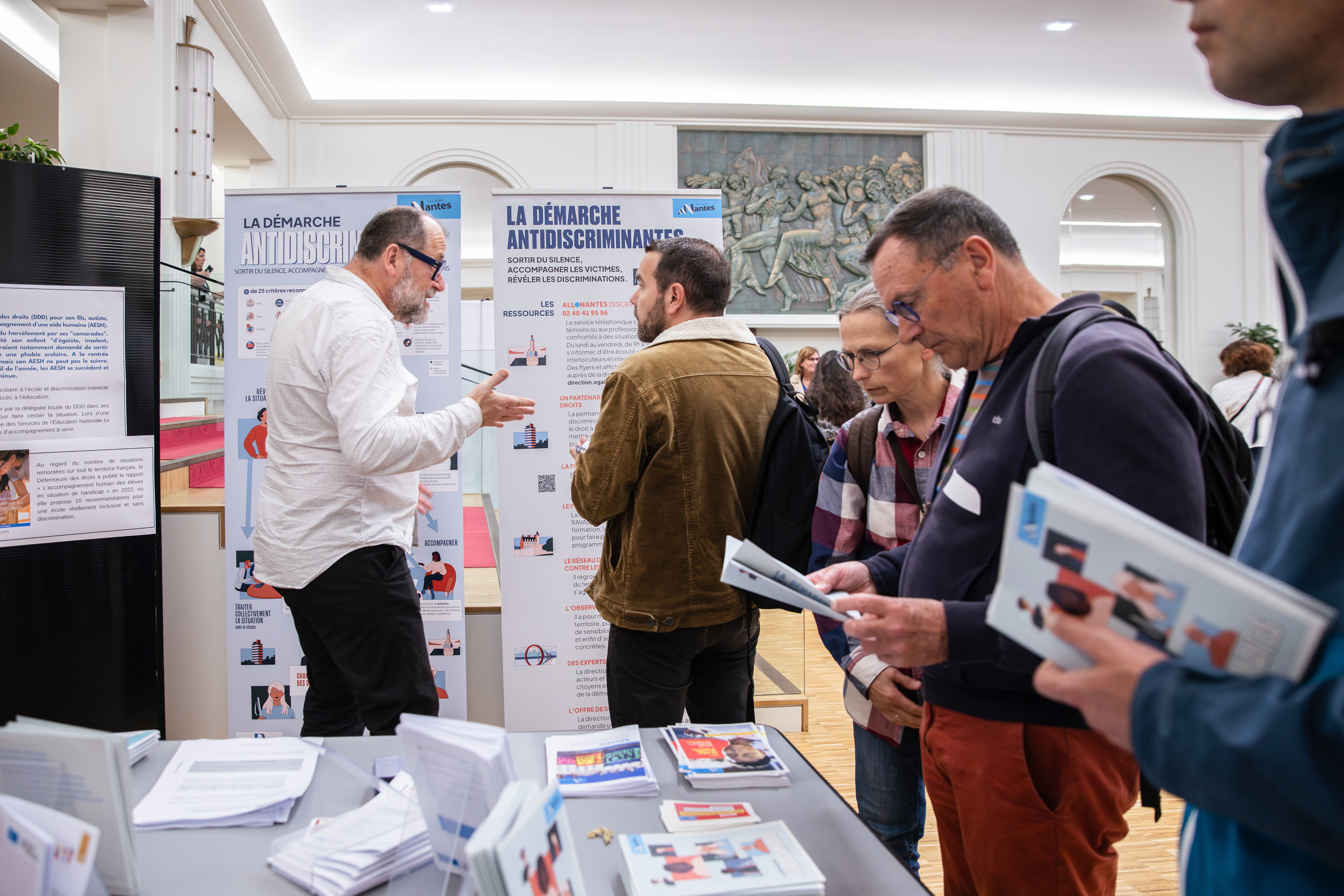 Des kakémonos de présentation de la démarche en fond, une table avec plusieurs prospectus et documents au premier plan, et plusieurs personnes discutant ou lisant des documents. 