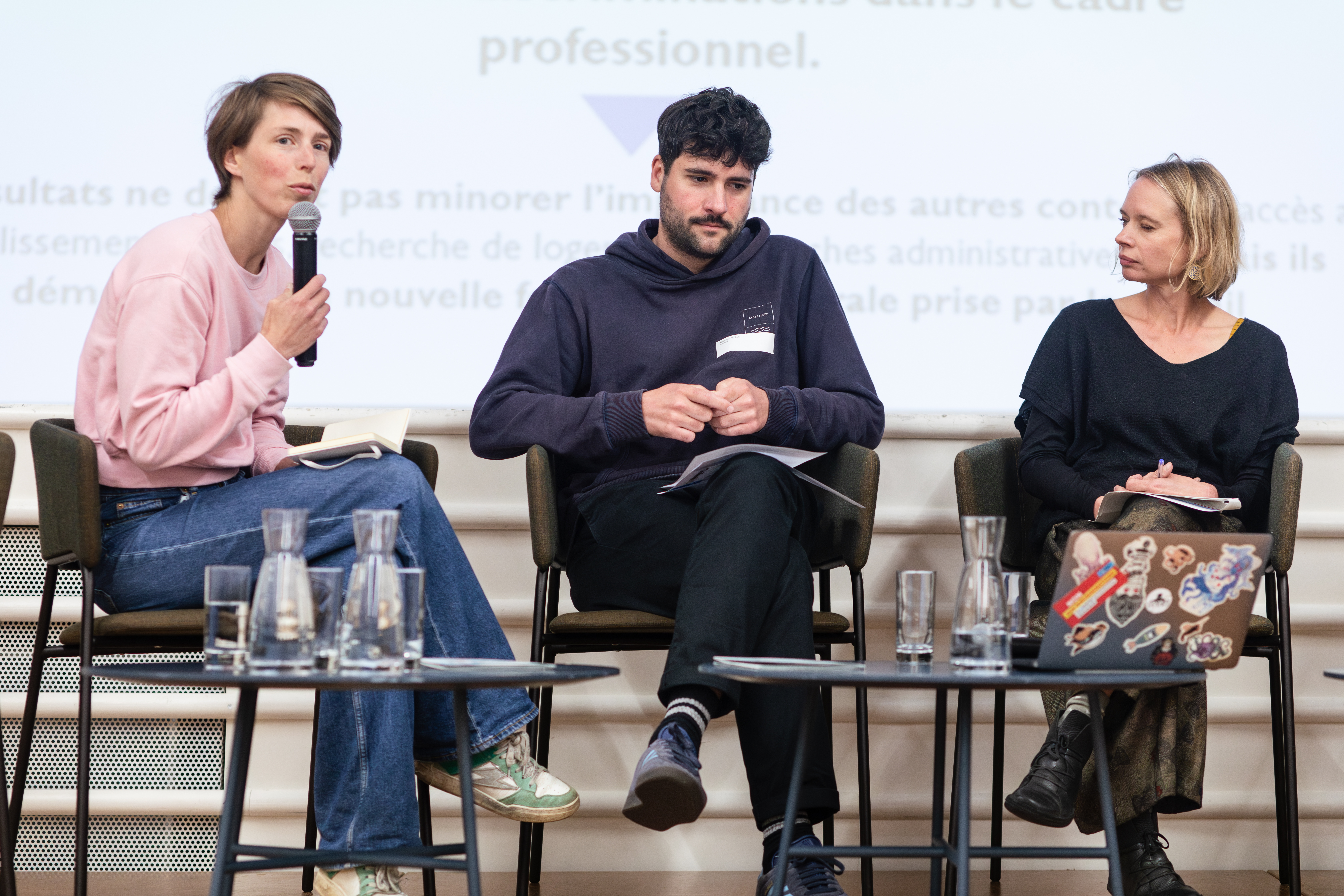 3 personnes sont en ligne sur la scène, 1 homme et deux femmes, dont l'une tient un micro et parle à au public que l'on ne voit pas sur l'image