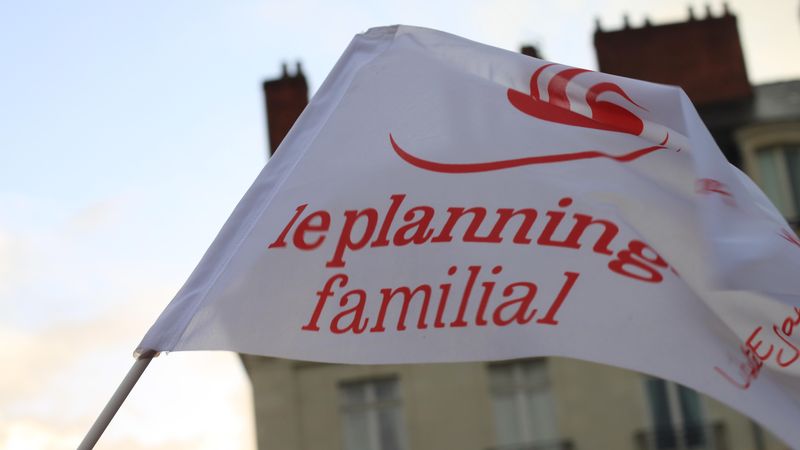 Drapeau du planning familial, texte rouge sur fond blanc, en arrière plan, le ciel bleu et un vieil immeuble