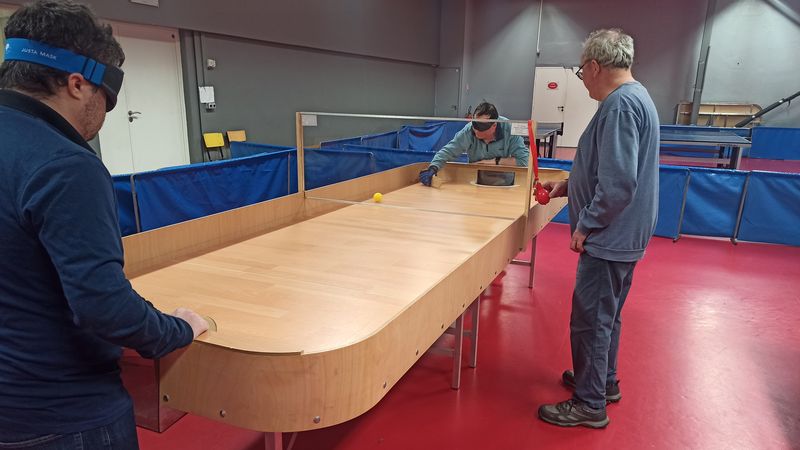 La photo montre deux hommes face à face à une table de showdown, portant des masques, un gant de protection tenant leur raquette de bois. L'un réalise un mouvement de revers et la balle jaune traverse la table sous l'oeil de l'arbitre. 