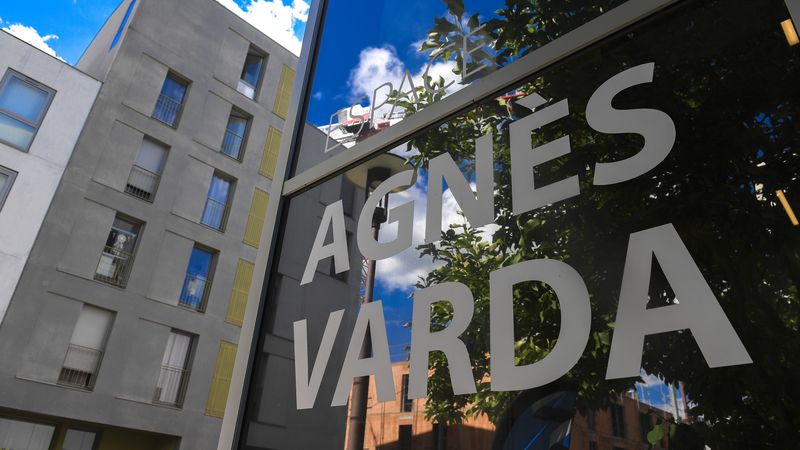 Photo de la vitrine de l'espace Agnès Varda ou figure le nom du batiment. En fond, un autre immeuble. 