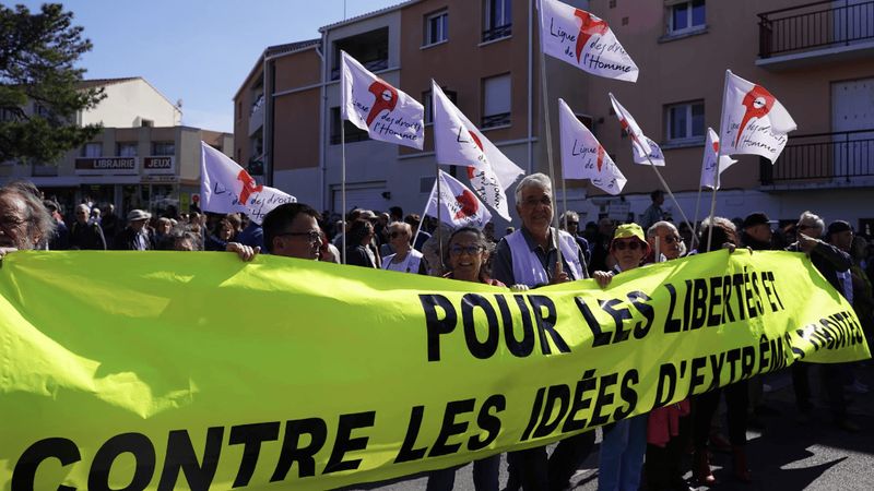 Photo d'une manifestation, ou des personnes portent des drapeaux de la ligue des droits de l'homme et une grande banderole jaune vif sur laquelle est inscrite 