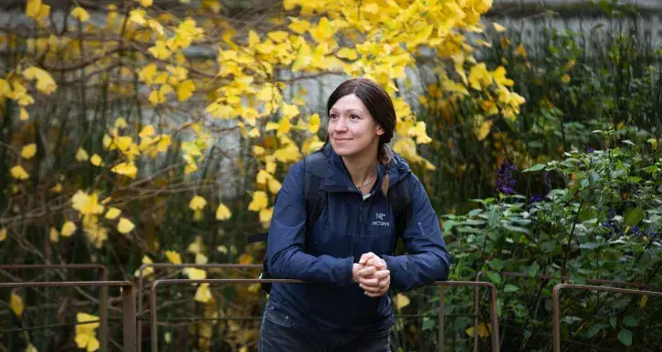 Ana Grau-Galofré souriante, en veste bleu foncé et sac à dos, s’appuie sur une barrière dans un jardin. Derrière elle, un arbre aux feuilles jaunes illumine la scène, créant une belle ambiance automnale.