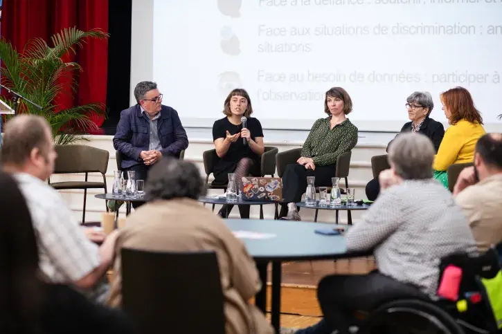 4 élus, 1 homme et deux femmes ainsi que l'animatrice et une sociologue sont assis en ligne sur la scène, sous le regard de plusieurs personnes assises à une table ronde au premier plan