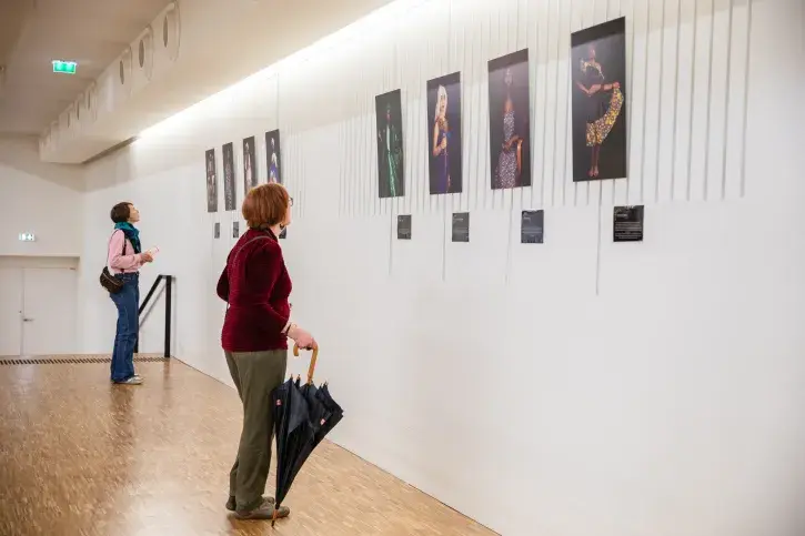 Plusieurs tableaux représentant des femmes sont accrochés sur un mur blanc. Devant, deux personnes sont en train de regarder les oeuvres. 
