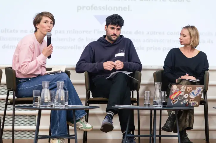 3 personnes sont en ligne sur la scène, 1 homme et deux femmes, dont l'une tient un micro et parle à au public que l'on ne voit pas sur l'image