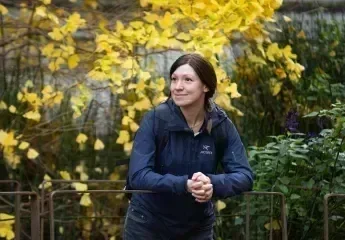 Ana Grau-Galofré souriante, en veste bleu foncé et sac à dos, s’appuie sur une barrière dans un jardin. Derrière elle, un arbre aux feuilles jaunes illumine la scène, créant une belle ambiance automnale.